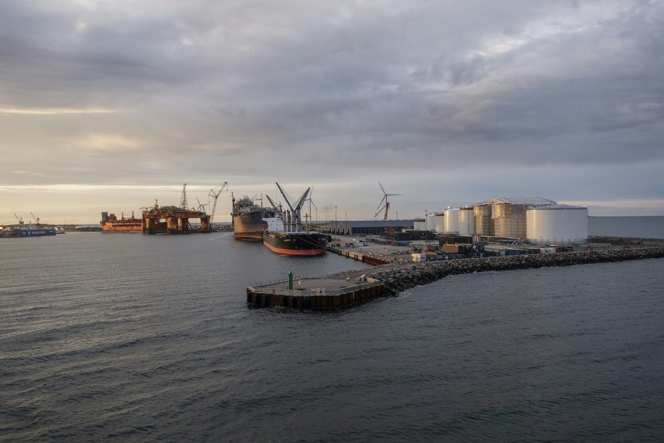 A wide industrial port and energy terminal, with an offshore platform, a moored vessel, storage tanks and wind turbines along the waterfront