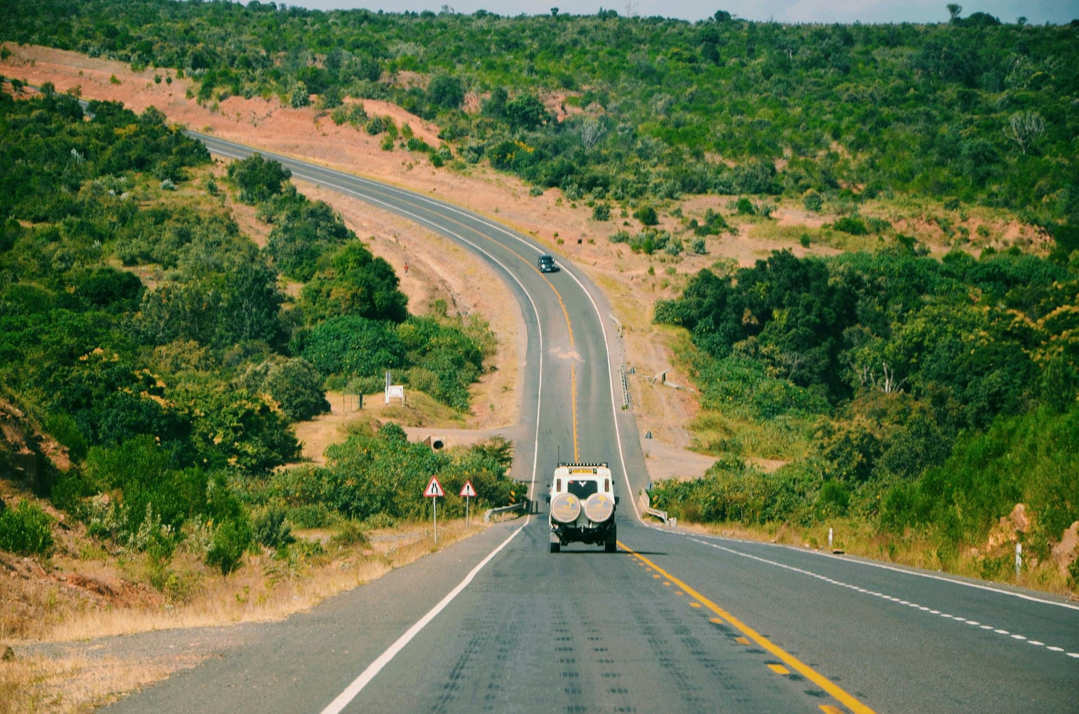 A two-lane highway winding through green hills, with a vehicle travelling the road toward a distant curve