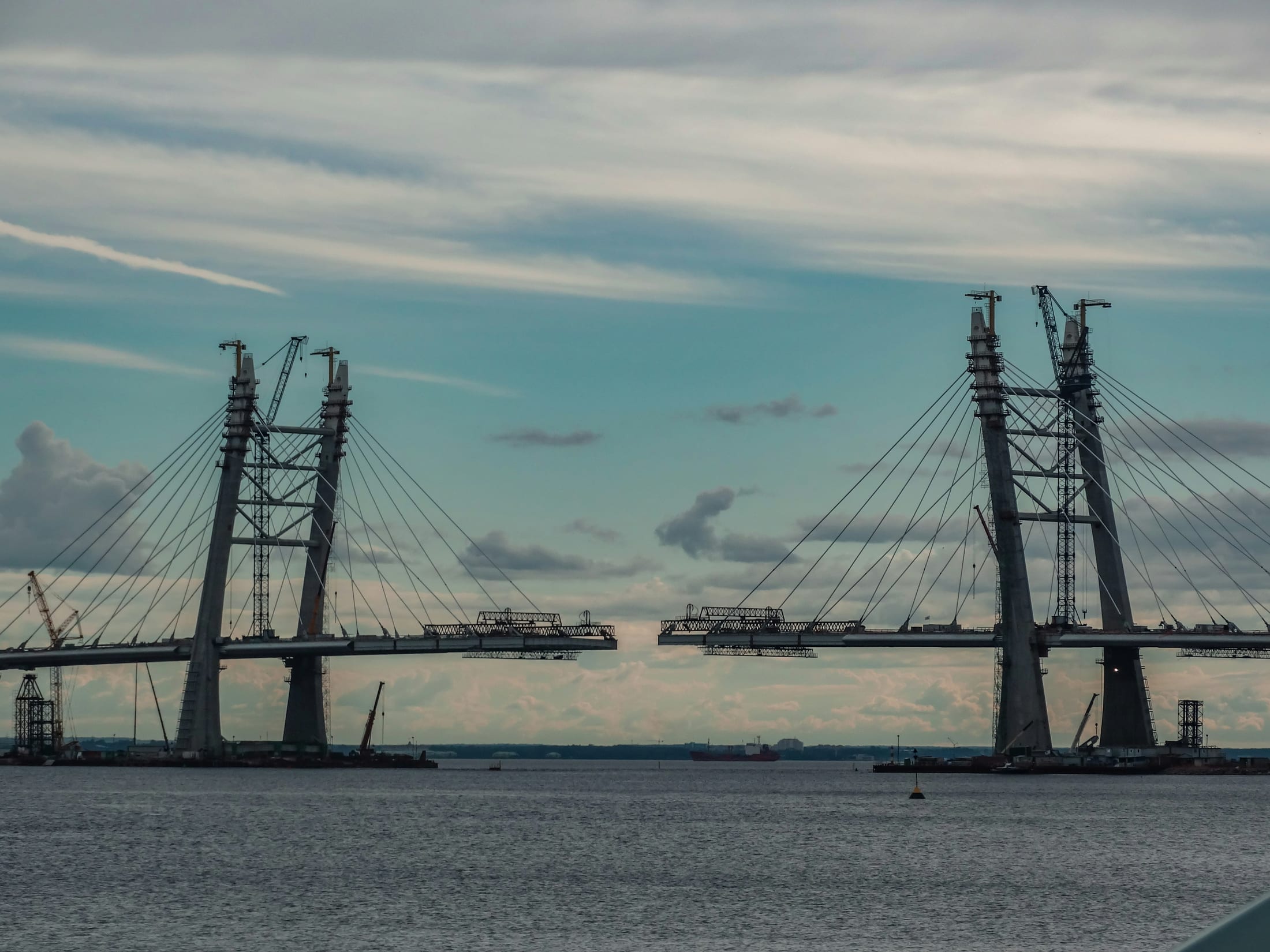 A cable-stayed bridge under construction, its two unfinished spans reaching toward each other across the water, with tower cranes and cabling visible against the sky