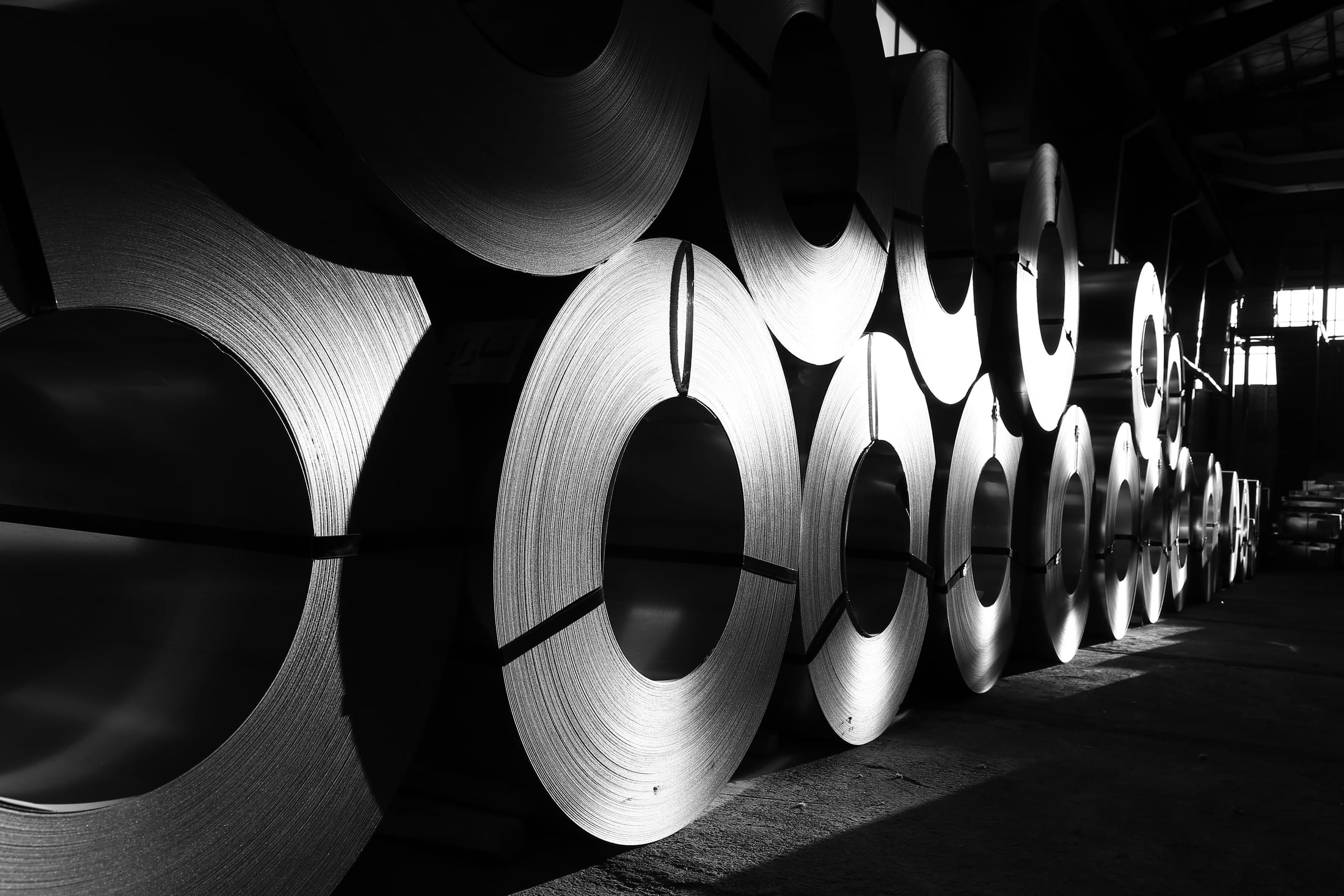 Rows of large rolled steel coils stacked in an industrial plant, lit from behind against a dark interior