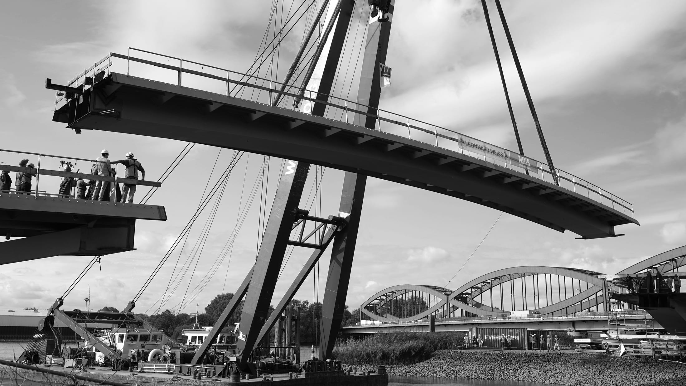 A suspended bridge span lifted into position by crane during construction, with workers overseeing the operation from an adjacent platform