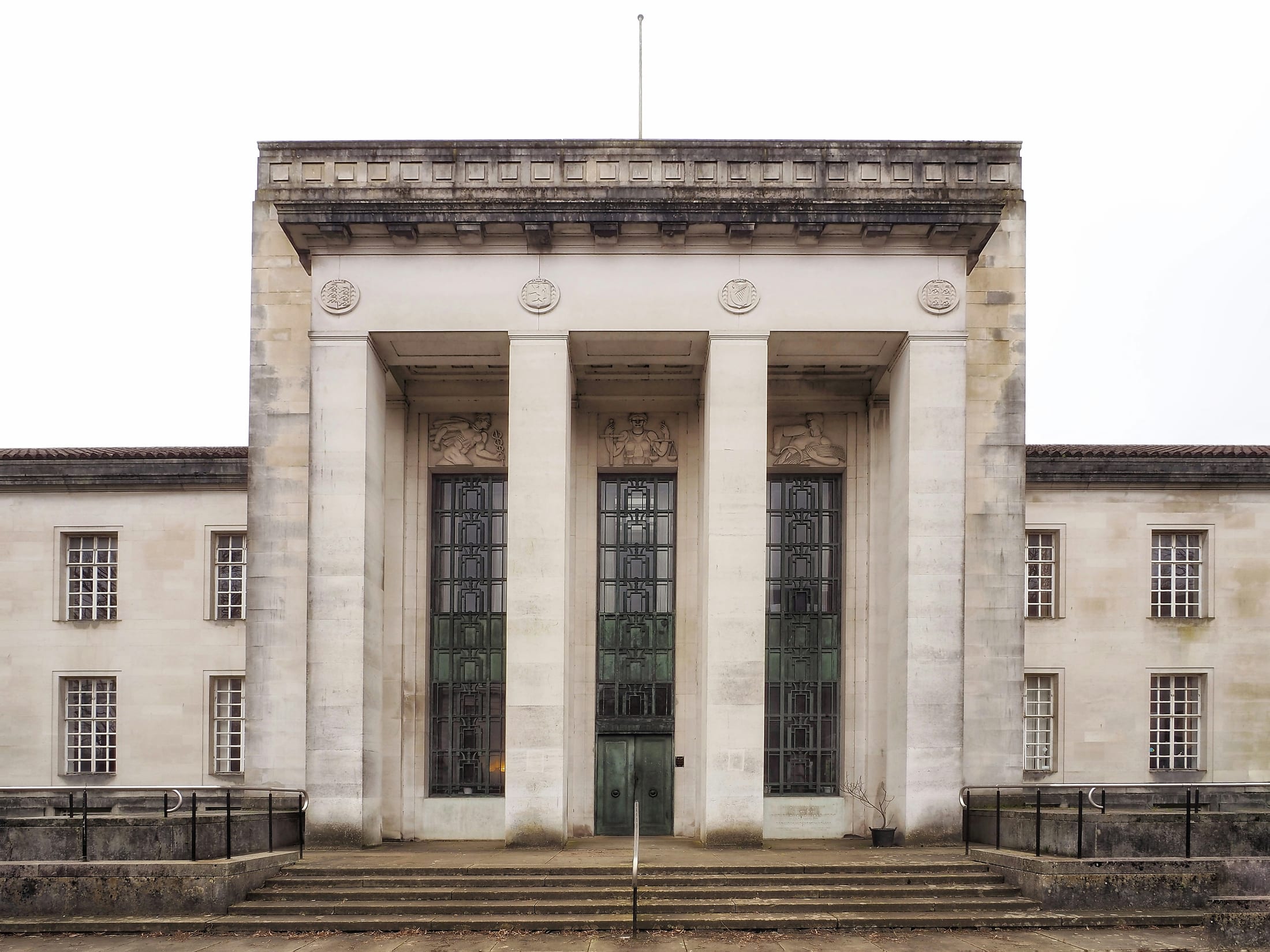 The symmetrical stone facade of an institutional building, with a colonnaded portico, carved relief panels and a flight of entrance steps
