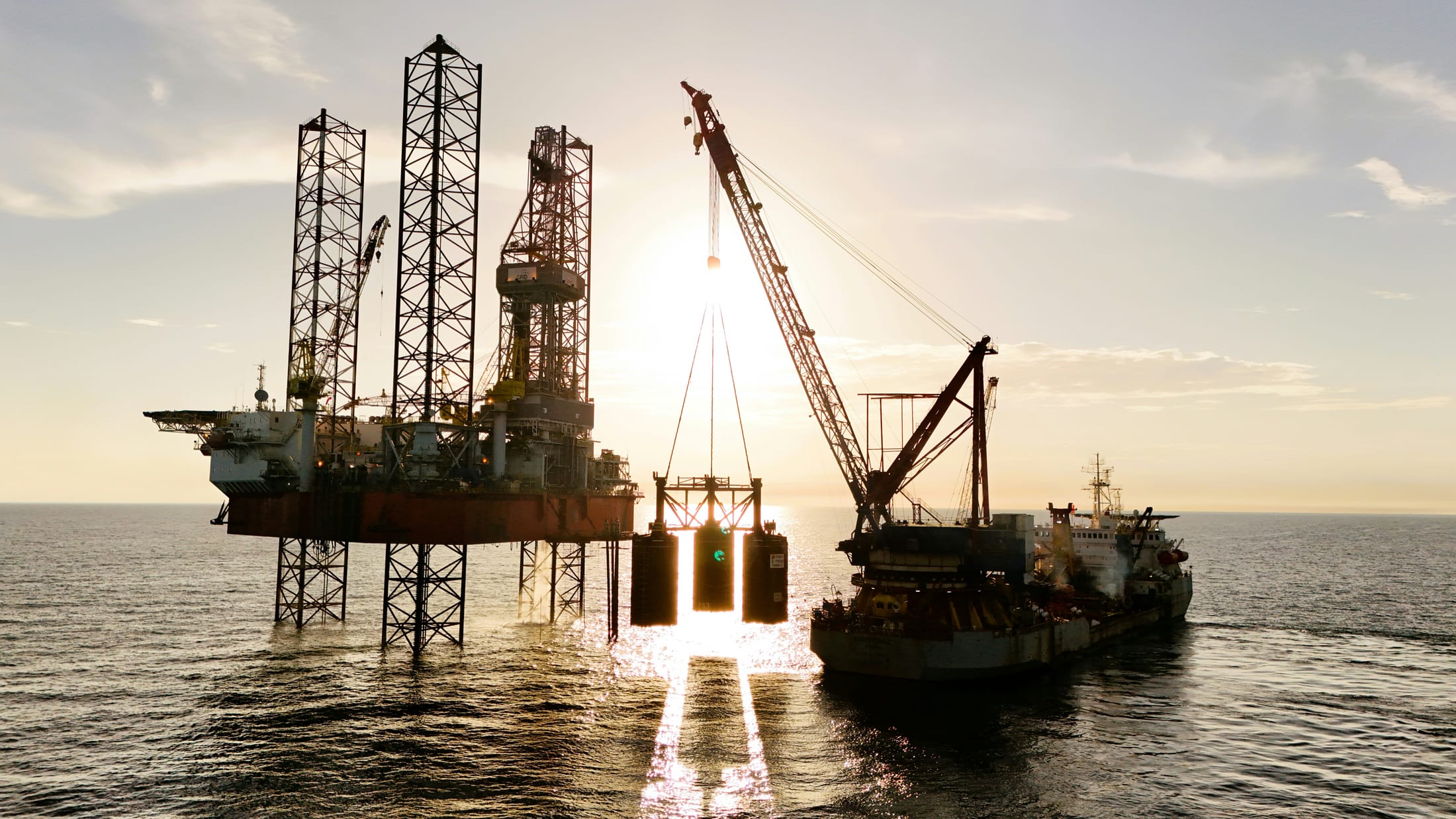 A jack-up rig beside a heavy-lift crane vessel lowering a suspended offshore structure into place, silhouetted against the setting sun