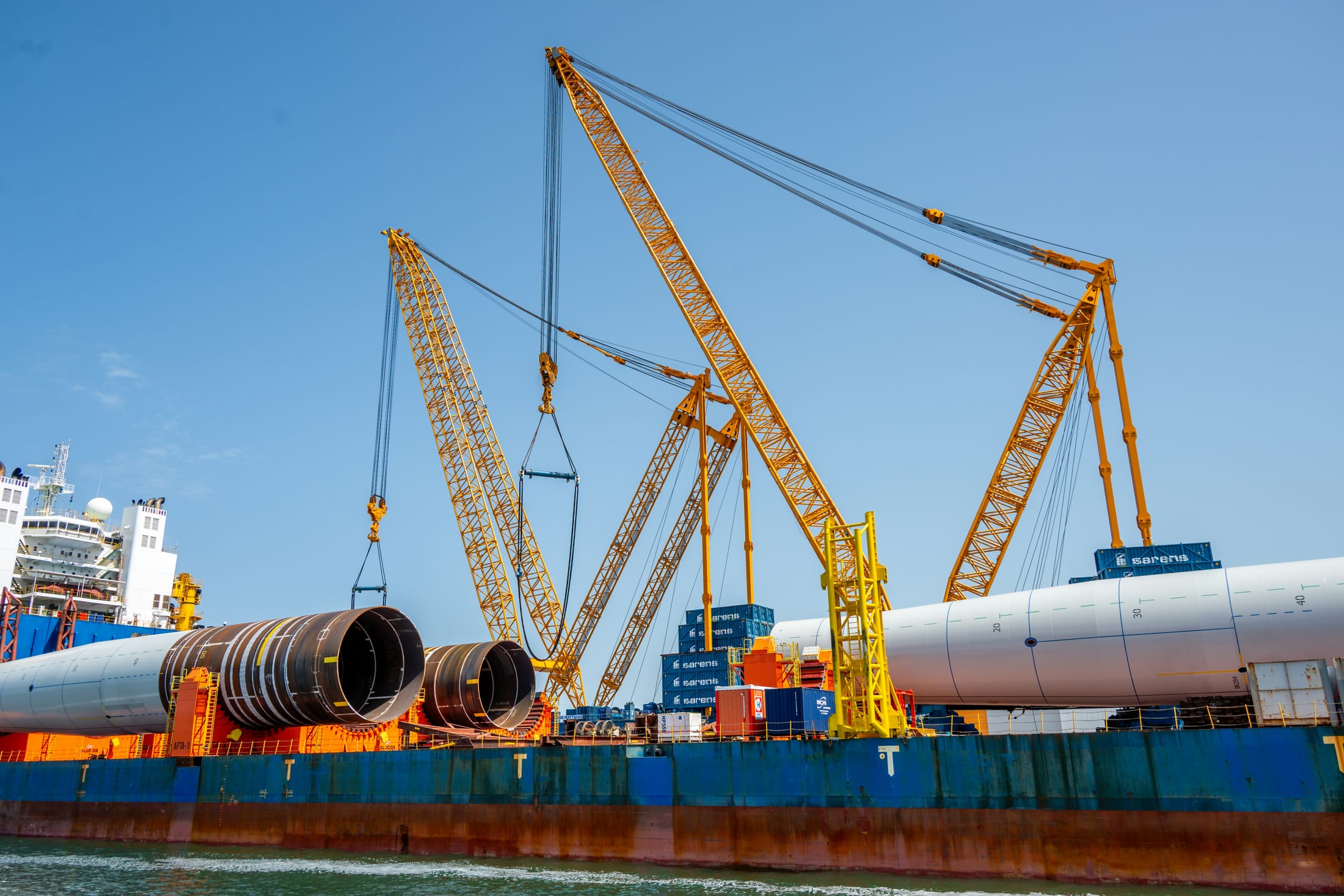 A heavy-lift barge with multiple cranes loading large fabricated steel pipe and tower sections for transport