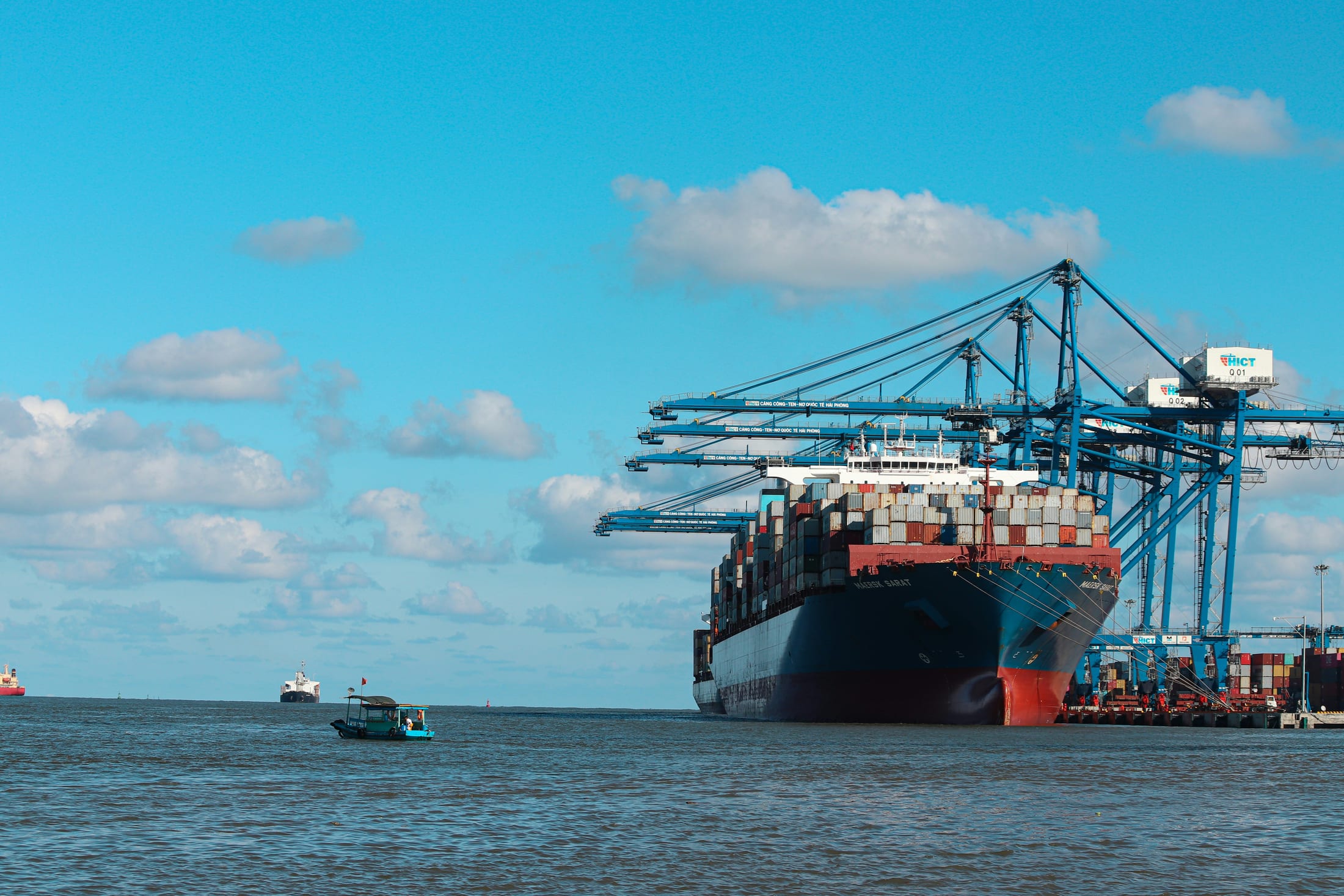 A loaded container vessel alongside gantry cranes at a working port, with a small boat passing in the foreground